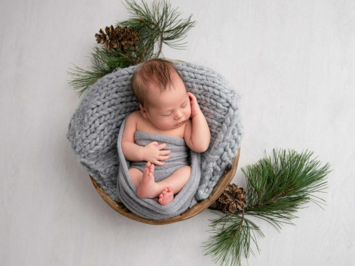 Vastasyntynyt poikavauva harmaassa kapalossa nukkumassa korissa kuusen oksien ympärillä