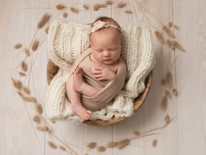 Vastasyntynyt vauva beige kapalossa ja kukka hiuspannassa nukkumassa ruskeassa korissa heinäpampula ympärillä