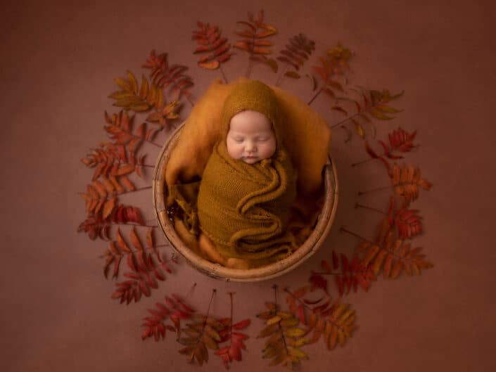 Vastasyntynyt vauva sinappivärisessä kapalossa ja myssyssä nukkumassa ruskeassa korissa syksylehtien ympärillä