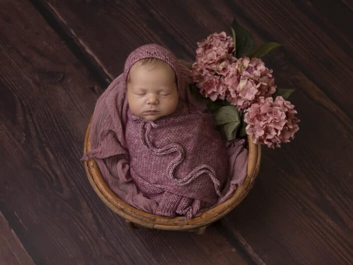 Vastasyntynyt vauva vaaleanpunaisessa kapalossa ja myssyssä nukkumassa korissa kukkien ympärillä