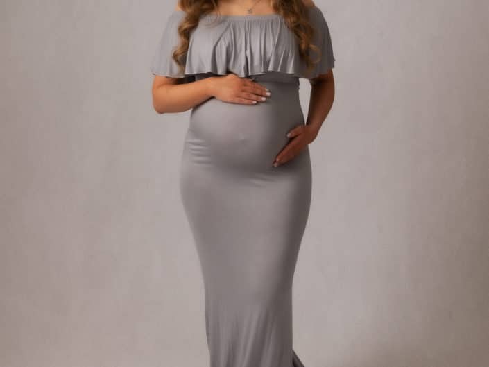 Raskaana oleva nainen harmaassa raskausmekossa vaalealla taustalla studiossa