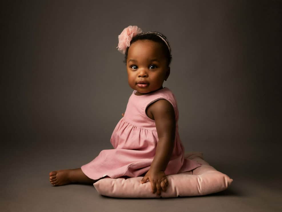 Dark skin one year old girl in a pink outfit sitting on a pink cushion