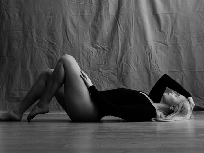 Nuori nainen mustassa asussa makaamassa lattialla, jalat koukussa, käsi koskettamassa hiuksia ja silmät olevia kiinni
