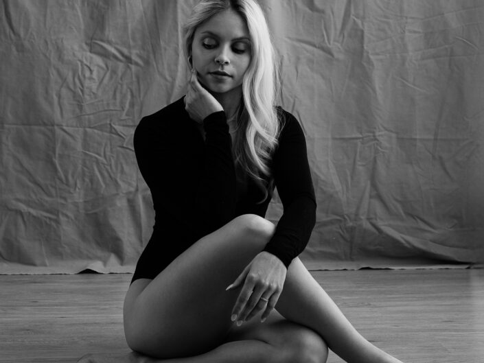 Nuori nainen mustassa bodyssa istumassa jalat ristissä, koskettamassa kasvonsa ja katsomassa alaspäin