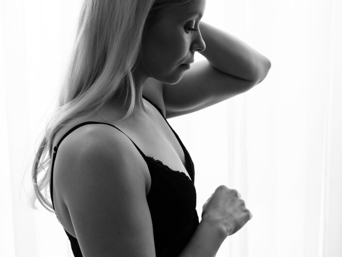 Nuori nainen mustassa asussa seisomassa valon edessä, koskettamassa kädellä hiuksia ja katsomassa alaspäin