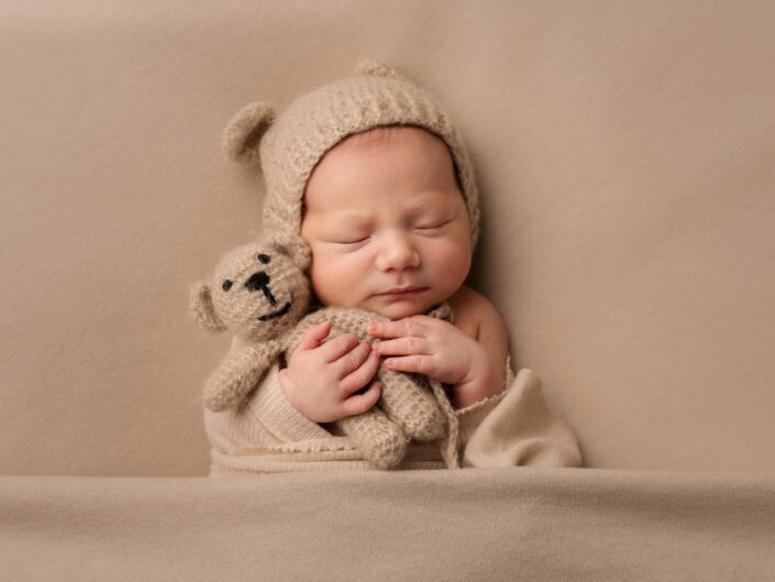 Vastasyntynyt vauva nukkuu käärittynä beigenväriseen peittoon, yllään neulottu hattu, jossa on karhun korvat ja jolla on virkattu nalle; pehmeä, yksivärinen beige tausta, Madlen Photography Jyväskylässä.