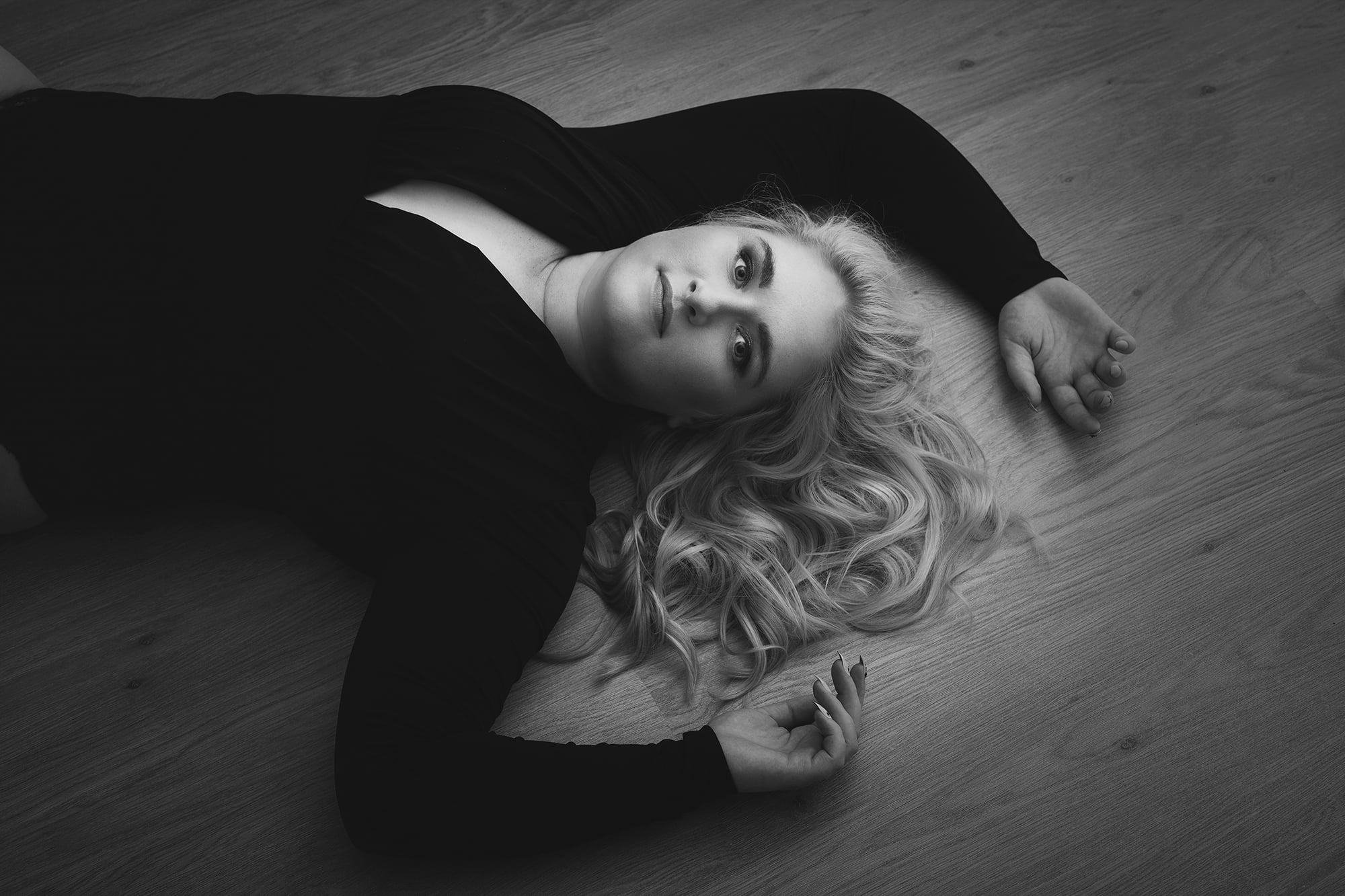 Madlen Photographyn elegantti mustavalkoinen budoaarivalokuvaus, jossa pitkä, aaltoileva hiuksinen nainen makaa kauniisti puulattialla. Hän käyttää tummaa, pitkähihaista toppia ja ojentaa kätensä katsellessaan ylöspäin ja huokuu intiimiä eleganssia.