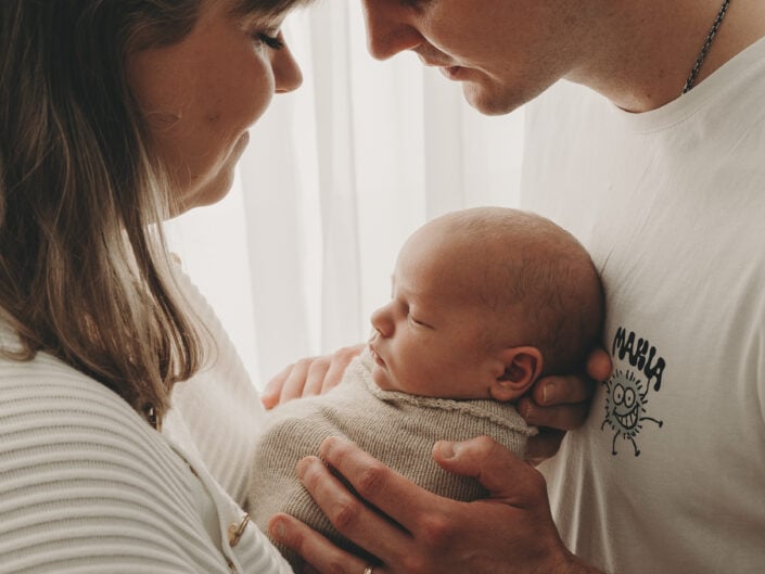 Pari Jyväskylässä, joka kehtaa nukkuvaa vauvaa beigenväriseen peittoon käärittynä, toisiaan päin ja hellästi koskettaa otsaa intiimin Madlen Photography -istunnon aikana lämpimässä, pehmeässä valaistuksessa.