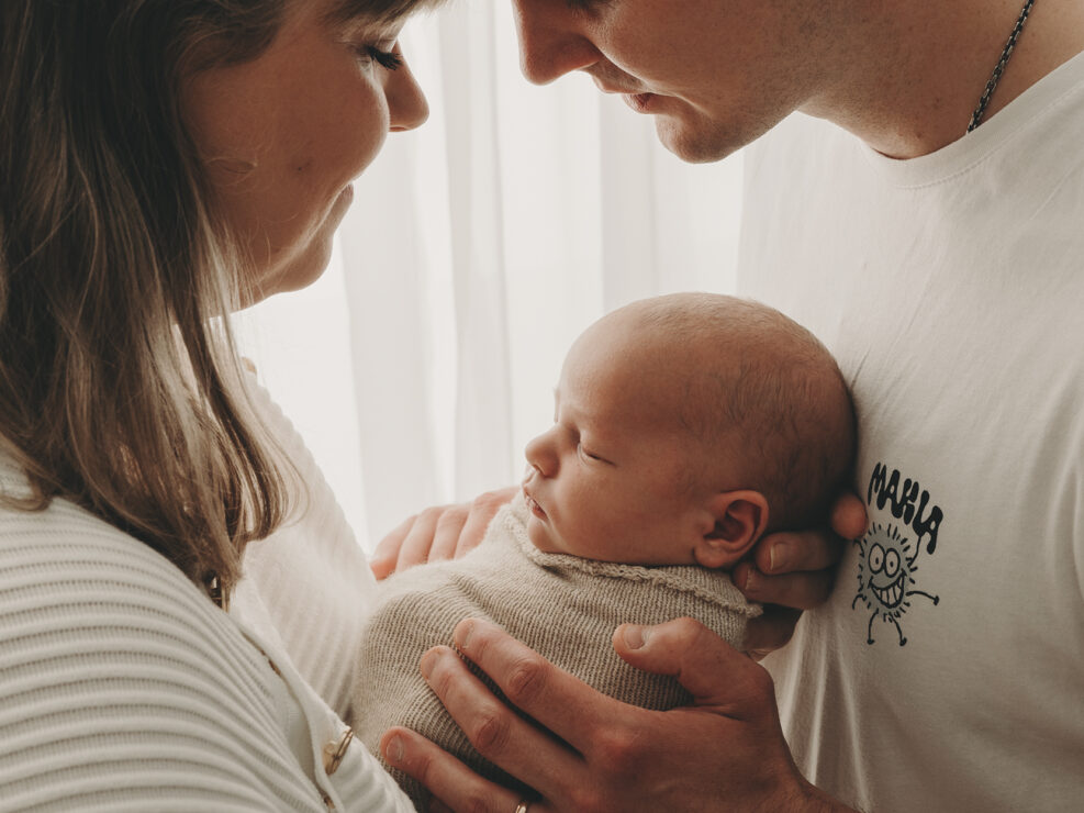 Pari Jyväskylässä, joka kehtaa nukkuvaa vauvaa beigenväriseen peittoon käärittynä, toisiaan päin ja hellästi koskettaa otsaa intiimin Madlen Photography -istunnon aikana lämpimässä, pehmeässä valaistuksessa.