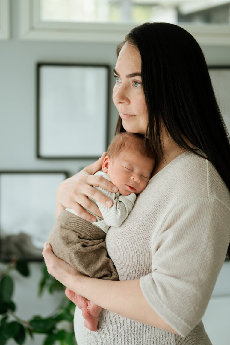 Äiti, jolla on pitkät tummat hiukset, halaa vastasyntynyttä vauvaa pehmeästi valaistussa Jyväskylän kodissa, Madlen Photography, takana kehystettyjä valokuvia ja kasvi.