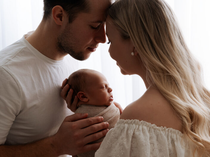 Vastasyntyneiden perhekuvaus Jyväskylässä: vanhemmat pitelevät vauvaa hellästi sylissä, otsat koskettavat, pehmeä luonnonvalo by Madlen Photography.