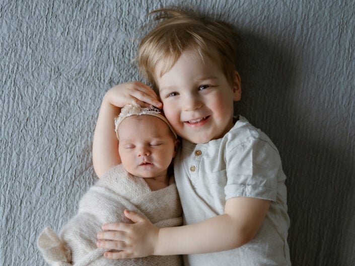 Madlen Photography, Jyväskylä: hymyilevä lapsi harmaalla huovalla halaamassa vastasyntynyttä vauvaa beigeen kääreeseen. Paikallinen perhevalokuvaus.