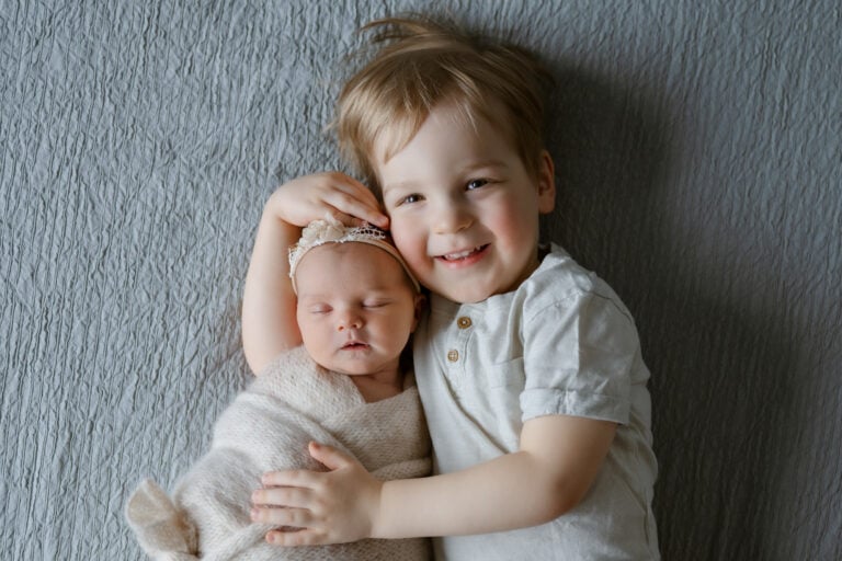 Madlen Photography, Jyväskylä: hymyilevä lapsi harmaalla huovalla halaamassa vastasyntynyttä vauvaa beigeen kääreeseen. Paikallinen perhevalokuvaus.
