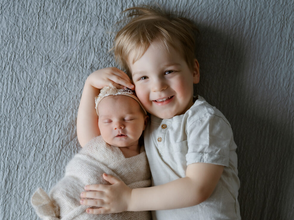Madlen Photography, Jyväskylä: hymyilevä lapsi harmaalla huovalla halaamassa vastasyntynyttä vauvaa beigeen kääreeseen. Paikallinen perhevalokuvaus.