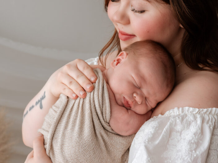 Äiti hymyilee hellästi ja pitelee nukkuvaa vauvaansa Jyväskylän studiossa by Madlen Photography, valkoinen mekko ja beige huopa.