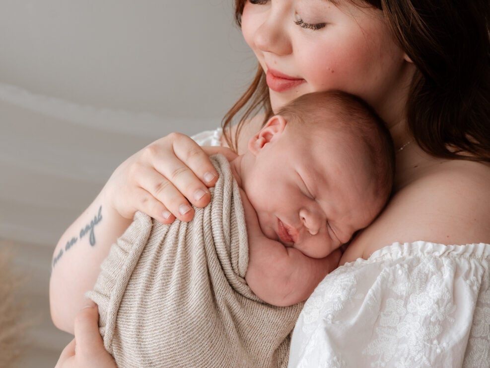 Äiti hymyilee hellästi ja pitelee nukkuvaa vauvaansa Jyväskylän studiossa by Madlen Photography, valkoinen mekko ja beige huopa.