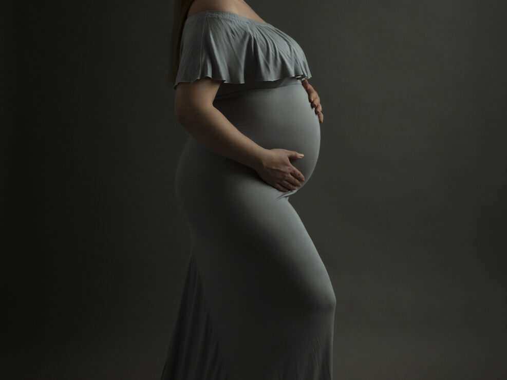 Madlen Photography:n ottama raskausvalokuva Jyväskylässä, jossa pitkä, olkapäät paljastava iltapuku päällään oleva raskaana oleva nainen seisoo profiilissa tummaa taustaa vasten, kädet vatsallaan ja katse eteenpäin suunnattuna.