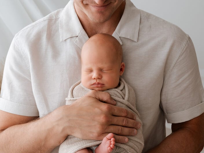 Isä vaaleassa paidassa pitelee hellästi sylissään nukkuvaa vastasyntynyttä, joka on kääritty beigeen peittoon. Madlen Photography on ikuistanut tämän hetken neutraalilla taustalla Jyväskylässä.
