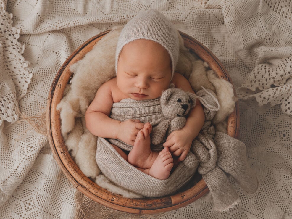 Madlen Photography:n vastasyntyneiden valokuvaus Jyväskylässä: rauhallisesti nukkuva vastasyntynyt vauva, kääritty beigeen kankaaseen pyöreän puisen kulhon sisällä, syleilemässä pientä neulottua nallekarhua kuvioidulla pitsipeitolla.