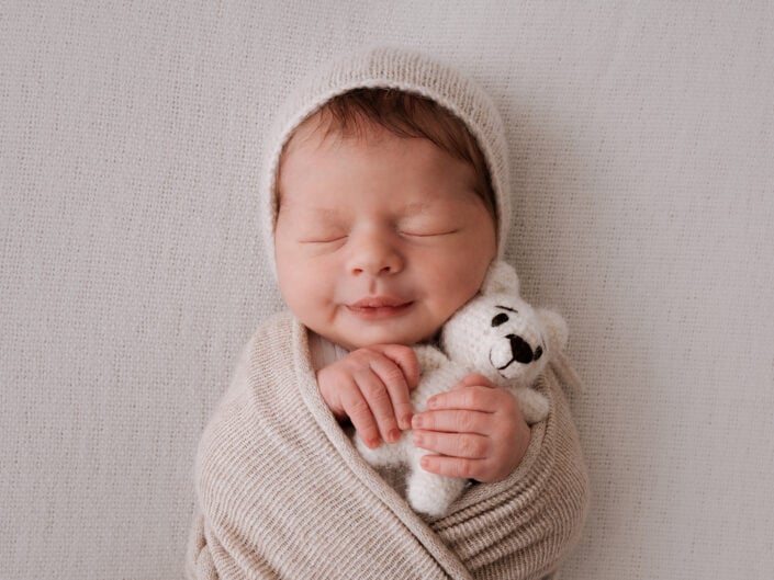 Beigeen peittoon ja myssyyn kääritty vastasyntynyt vauva, joka nukkuu rauhallisesti ja hymyilee pientä valkoista neulottua karhua sylissään; kuvannut Madlen Photography pehmeällä alustalla Jyväskylässä.